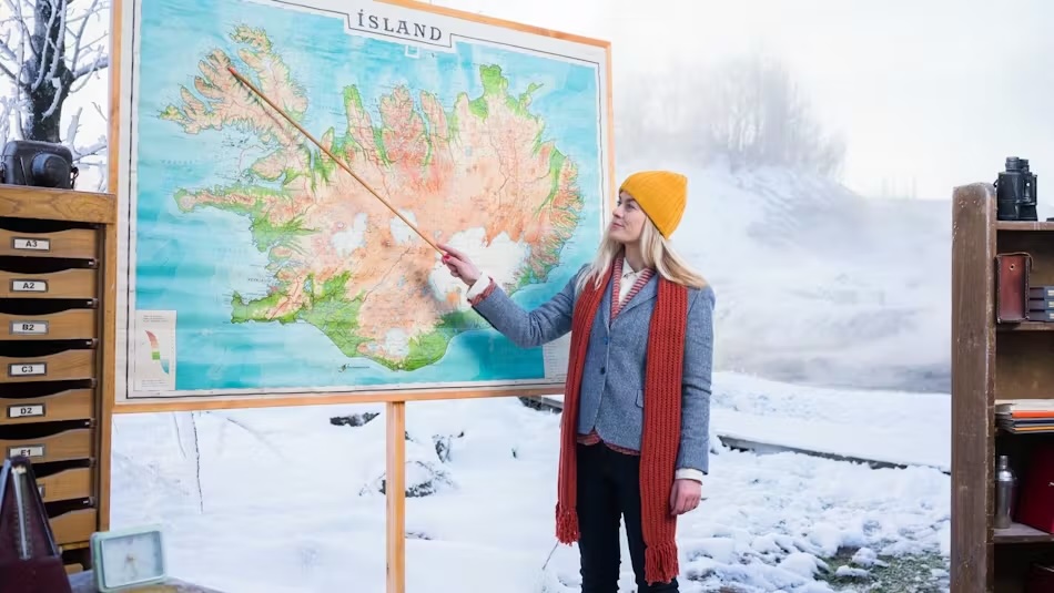 A woman wearing a yellow beanie, a blue coat, and an orange scarf is standing in a snowy outdoor setting. She is pointing to a large map of Iceland with a wooden pointer. The map is mounted on a stand, and there is a wooden shelf with labeled drawers on the left side of the image. On the right side, there is a wooden shelf with various items, including a camera. The background features snow-covered trees and a misty atmosphere.