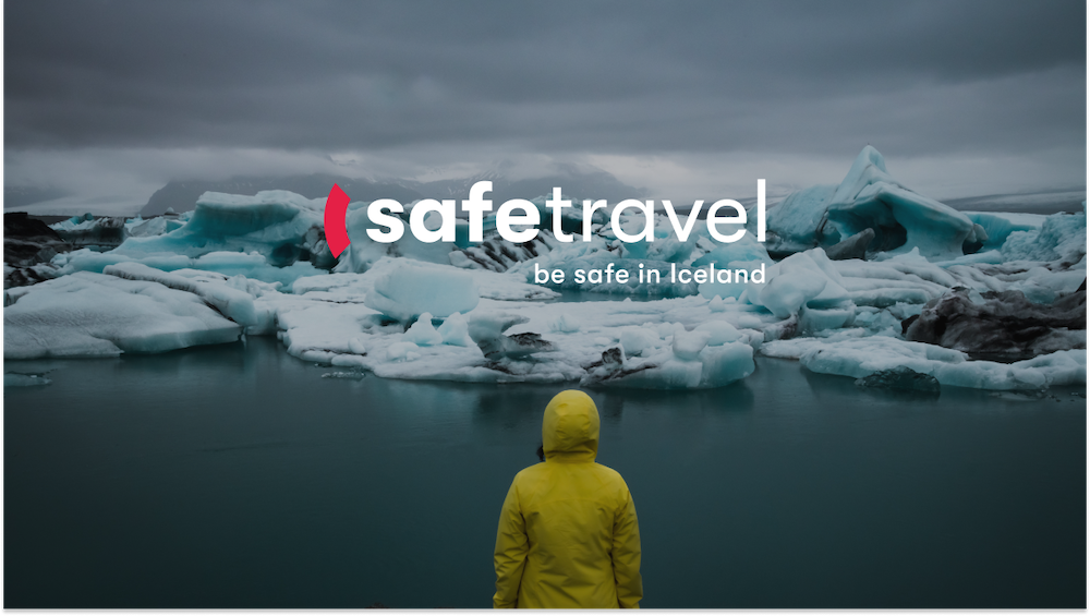 “An individual in a yellow raincoat stands facing a serene, icy landscape with large ice formations and a calm body of water. The sky is overcast with dark clouds. Overlaid text reads ‘safetravel - be safe in Iceland’ with a red partial circle symbol to the left of ‘safe’.”