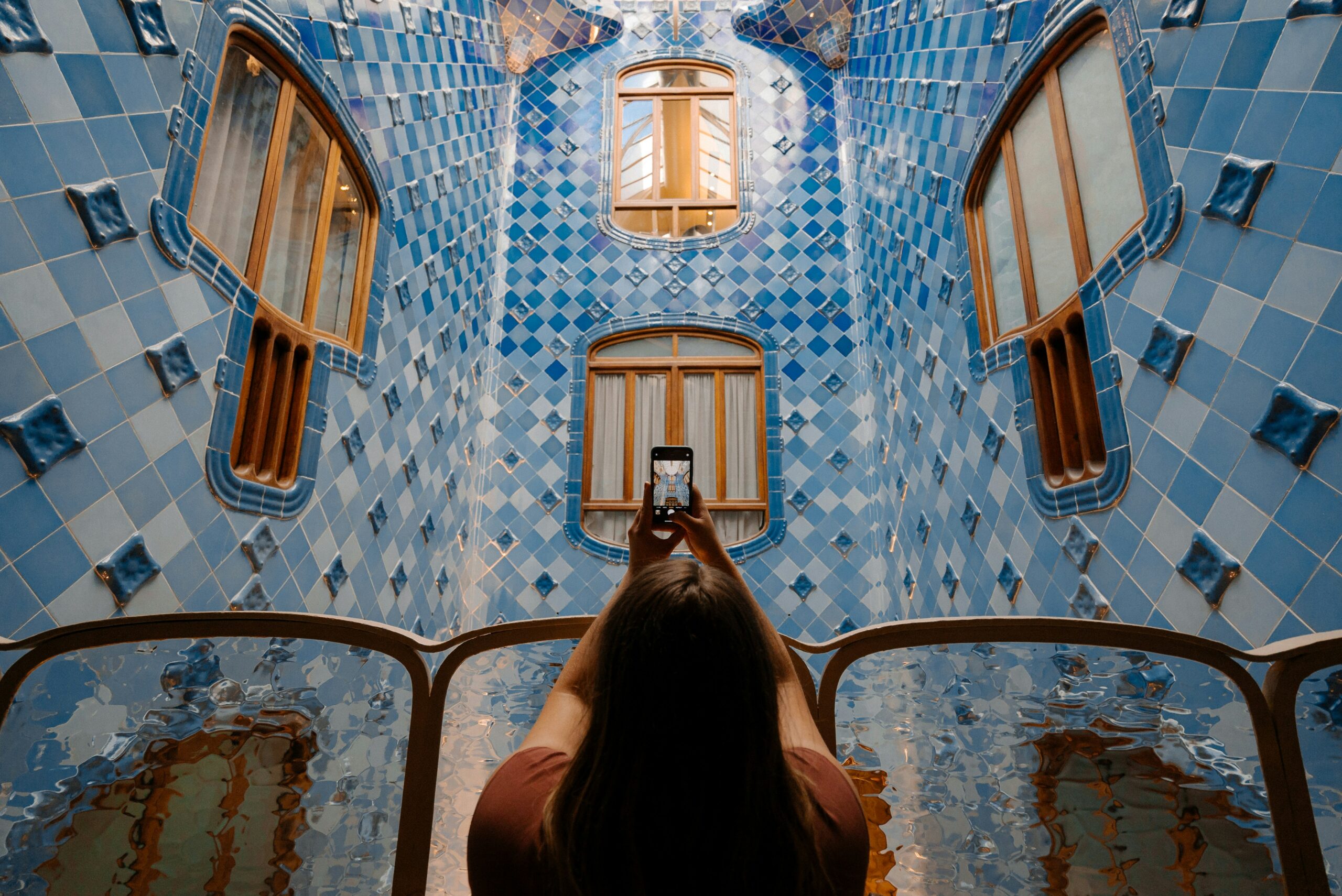 A person stands inside a blue-tiled room, holding up a smartphone to take a picture of the intricate architectural design. The room features various shades of blue tiles and curved wooden windows.