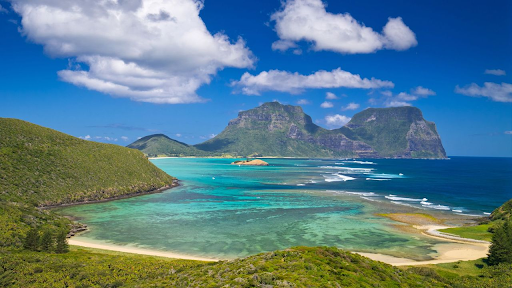 A scenic aerial view of Lord Howe Island, Australia, showcasing a pristine beach, turquoise lagoon, lush green hills, and towering mountains under a clear blue sky.