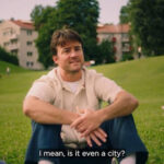 A man sitting on a grassy field, wearing a light-colored shirt and blue pants, smiling as he casually gestures with his hands. In the background, people are relaxing in the park, with buildings and trees visible. The caption at the bottom reads, "I mean, is it even a city?"