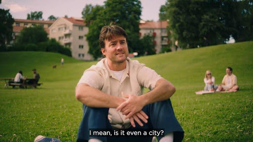 A man sitting on a grassy field, wearing a light-colored shirt and blue pants, smiling as he casually gestures with his hands. In the background, people are relaxing in the park, with buildings and trees visible. The caption at the bottom reads, "I mean, is it even a city?"