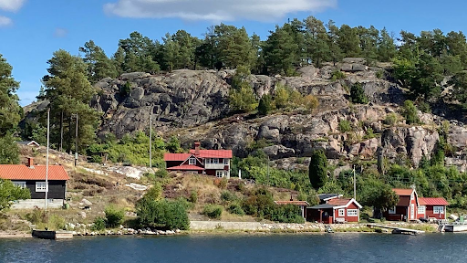 A scenic view of a rocky hillside by the water, featuring several small, red and black wooden houses surrounded by trees and greenery under a clear blue sky.