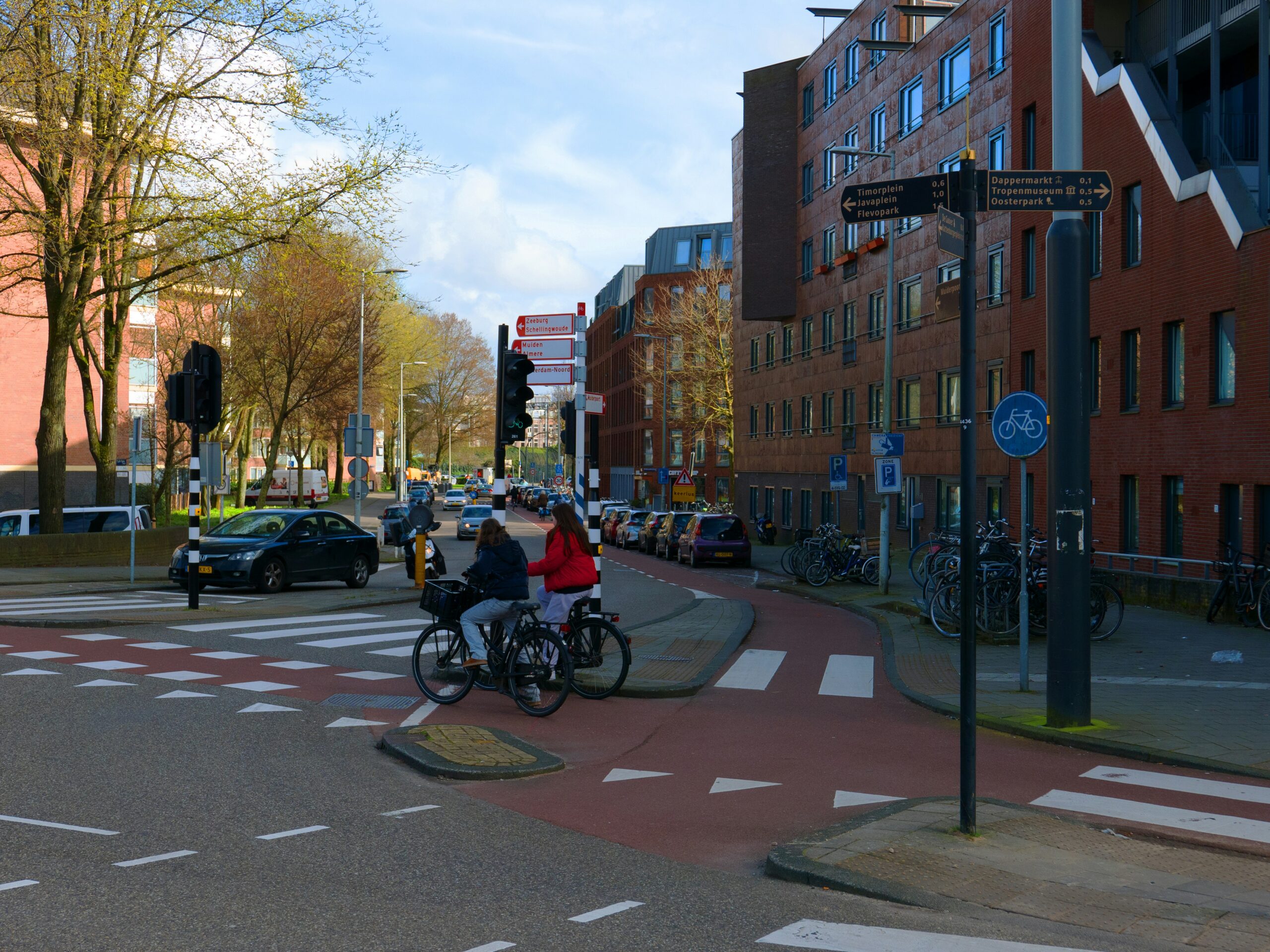 A city intersection with a designated red bike lane in the Netherlands. Two cyclists wait at a crosswalk, surrounded by buildings, trees, parked cars, and traffic signs. The bike path is clearly marked, and there are separate traffic signals for cyclists.