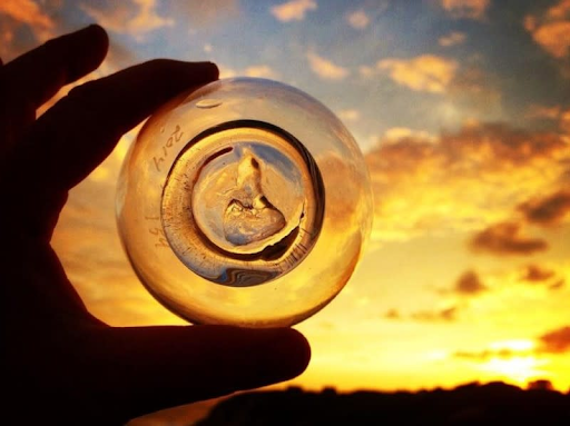 A hand holds a circular piece of glass against a vibrant sunset sky, capturing the warm hues of orange and yellow. The glass appears to contain a textured design that resembles a wolf's head.