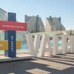 A colorful display structure with intersecting rectangular panels stands in front of modern architecture and a water feature in Valencia. The panels read "VALENCIA 2022," "EUROPEAN CAPITAL," "OF SMART TOURISM," each in different colors and fonts, celebrating Valencia's recognition as the European Capital of Smart Tourism.