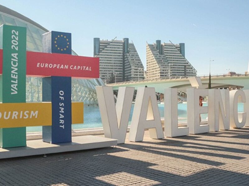 A colorful display structure with intersecting rectangular panels stands in front of modern architecture and a water feature in Valencia. The panels read "VALENCIA 2022," "EUROPEAN CAPITAL," "OF SMART TOURISM," each in different colors and fonts, celebrating Valencia's recognition as the European Capital of Smart Tourism.