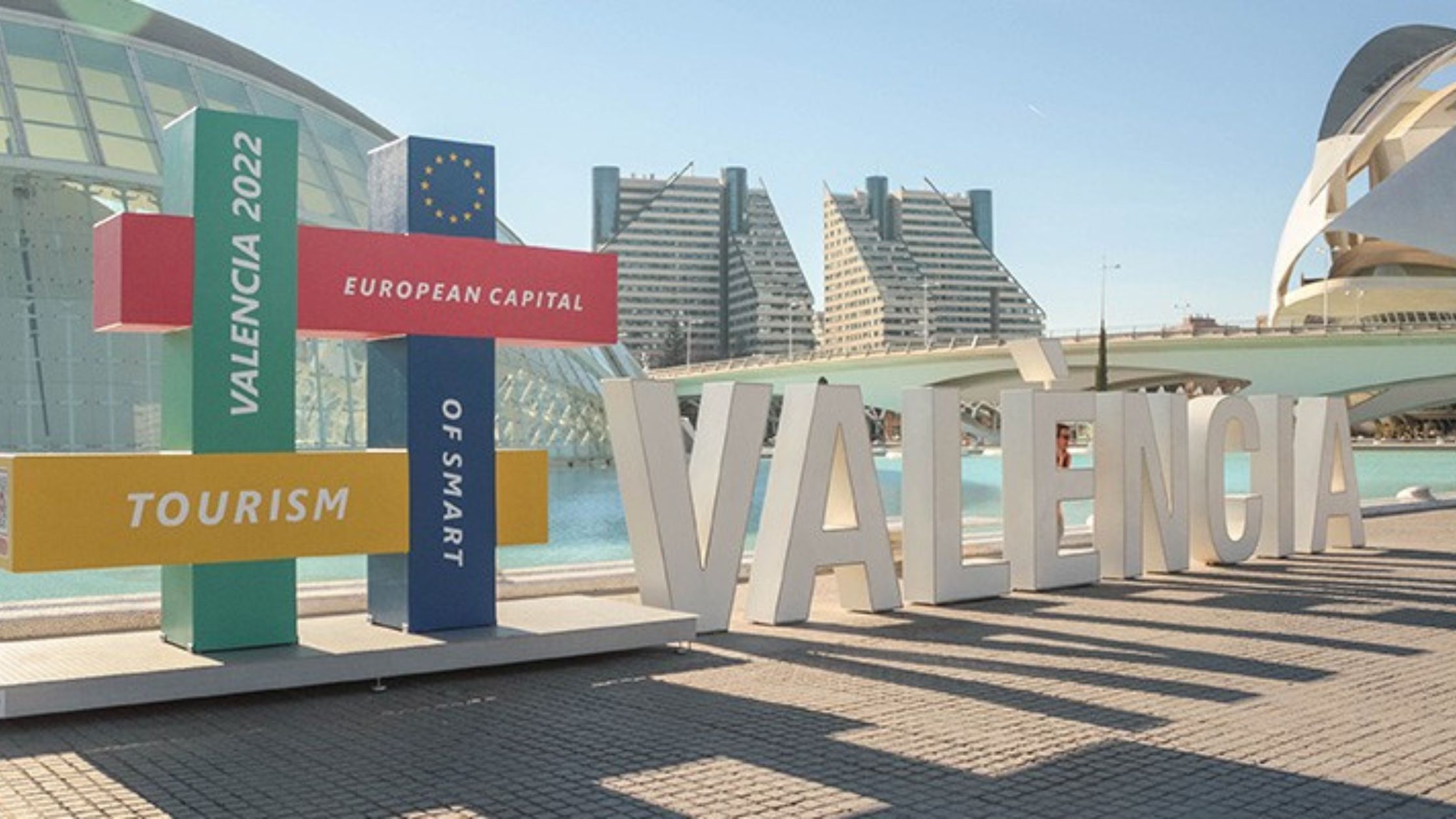 A colorful display structure with intersecting rectangular panels stands in front of modern architecture and a water feature in Valencia. The panels read "VALENCIA 2022," "EUROPEAN CAPITAL," "OF SMART TOURISM," each in different colors and fonts, celebrating Valencia's recognition as the European Capital of Smart Tourism.