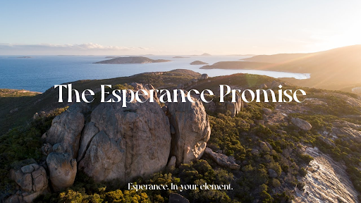 A scenic aerial view of Esperance, Australia, featuring rugged rock formations, lush green vegetation, and the deep blue ocean stretching toward the horizon. The golden light of the setting sun casts a warm glow over the landscape. White text in the center reads, "The Esperance Promise," with a tagline below stating, "Esperance. In your element."