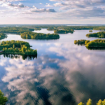 A stunning aerial view of a lake in Finland, featuring numerous small, tree-covered islands surrounded by calm, reflective waters. The sky is dotted with soft clouds, whose reflections create a mirror-like effect on the lake's surface. The lush greenery contrasts beautifully with the deep blue water, highlighting the serene natural beauty of the region.