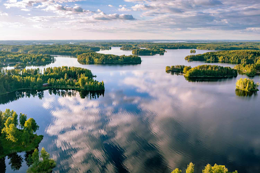 A stunning aerial view of a lake in Finland, featuring numerous small, tree-covered islands surrounded by calm, reflective waters. The sky is dotted with soft clouds, whose reflections create a mirror-like effect on the lake's surface. The lush greenery contrasts beautifully with the deep blue water, highlighting the serene natural beauty of the region.