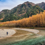 A person stands near the edge of a winding, crystal-clear river surrounded by a landscape of golden autumn trees and towering mountains in the background. The sky is a mix of blue and soft clouds, adding depth to the serene natural setting.