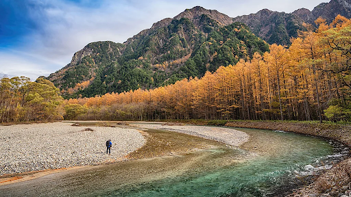 A person stands near the edge of a winding, crystal-clear river surrounded by a landscape of golden autumn trees and towering mountains in the background. The sky is a mix of blue and soft clouds, adding depth to the serene natural setting.