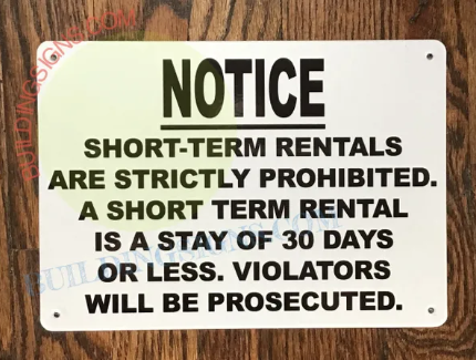 A white rectangular sign with black lettering stating "NOTICE SHORT-TERM RENTALS ARE STRICTLY PROHIBITED. A SHORT TERM RENTAL IS A STAY OF 30 DAYS OR LESS. VIOLATORS WILL BE PROSECUTED." The sign is affixed to a wooden surface.