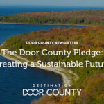 Aerial view of a forested shoreline in Door County, Wisconsin, with calm blue water and autumn foliage, overlaid with the text “The Door County Pledge: Creating a Sustainable Future.”