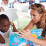 A woman reads a children’s book titled “Sharing a Shell” to a young boy at a table, where other books and papers are scattered. The boy listens attentively with his head resting on his hand. The background suggests a community or outdoor event with other people and chairs visible.
