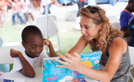 A woman reads a children’s book titled “Sharing a Shell” to a young boy at a table, where other books and papers are scattered. The boy listens attentively with his head resting on his hand. The background suggests a community or outdoor event with other people and chairs visible.