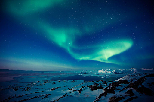 A stunning view of the Northern Lights (Aurora Borealis) illuminating the night sky with vibrant green and blue hues. The lights stretch across the sky, creating a mesmerizing display above a snowy, mountainous landscape. The stars are visible in the background, adding to the beauty of the scene.
