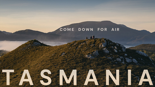A scenic view of hikers standing atop a grassy, rocky hill in Tasmania, with misty valleys and mountain ranges in the background. The text “Come Down for Air” appears above the hikers, and “TASMANIA” is boldly displayed at the bottom.