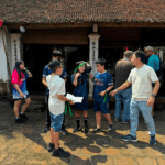 A group of children and a guide engage in a gamified heritage tour at an ancient village in Vietnam, standing in front of a traditional tiled-roof building.