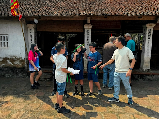 A group of children and a guide engage in a gamified heritage tour at an ancient village in Vietnam, standing in front of a traditional tiled-roof building.