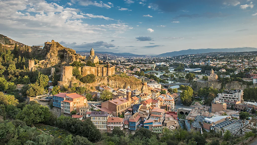 Panoramic view of Tbilisi, Georgia, featuring historic Narikala Fortress, colorful houses with balconies, and the sprawling cityscape under a partly cloudy sky.