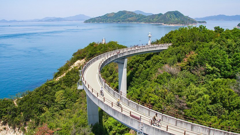 Cyclists ride along a scenic elevated bike path on Japan’s Shimanami Kaido, surrounded by lush greenery and overlooking the calm blue waters of the Seto Inland Sea.