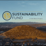 Aerial view of Pilot Butte in Bend, Oregon, surrounded by neighborhoods and forests under a clear sky. The logo for the Bend Sustainability Fund appears in the sky, with the tagline “A Visit Bend Project.”