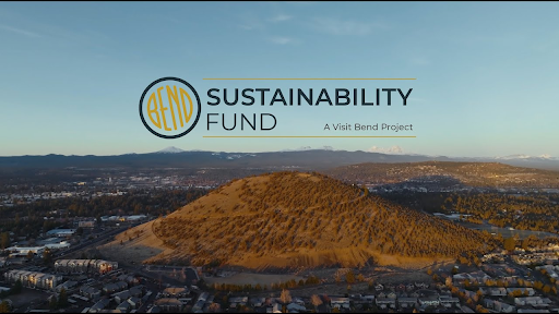 Aerial view of Pilot Butte in Bend, Oregon, surrounded by neighborhoods and forests under a clear sky. The logo for the Bend Sustainability Fund appears in the sky, with the tagline “A Visit Bend Project.”