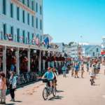 A lively street scene on Mackinac Island with people walking, biking, and shopping along storefronts decorated with hanging flowers and flags.