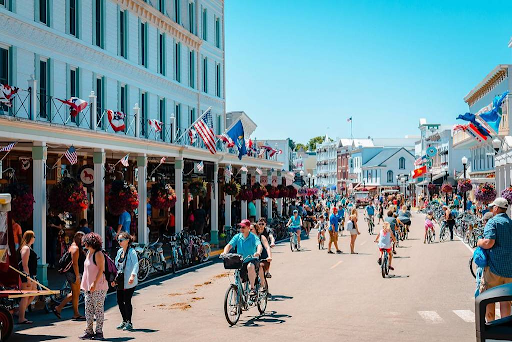 A lively street scene on Mackinac Island with people walking, biking, and shopping along storefronts decorated with hanging flowers and flags.