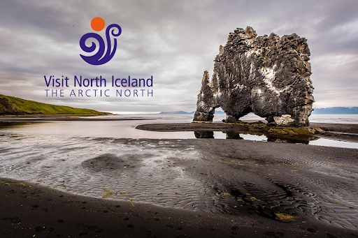 A dramatic coastal rock formation known as Hvitserkur rises from the black sand beach in North Iceland, with cloudy skies and distant mountains in the background. A logo reading “Visit North Iceland – The Arctic North” is placed on the image.