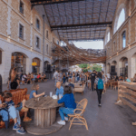 People gather in a lively open-air market hall in Bordeaux, France, sitting at wooden tables and walking between historic stone buildings with shops and restaurants.