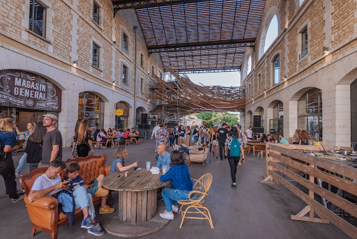 People gather in a lively open-air market hall in Bordeaux, France, sitting at wooden tables and walking between historic stone buildings with shops and restaurants.