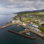Aerial view of a coastal town in North Iceland with a small harbor, fishing boats, colorful houses, and green hills under a cloudy sky.