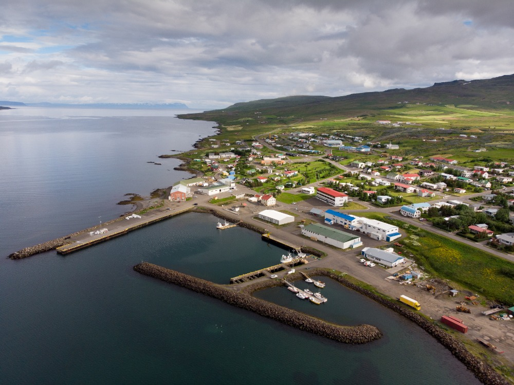 Aerial view of a coastal town in North Iceland with a small harbor, fishing boats, colorful houses, and green hills under a cloudy sky.