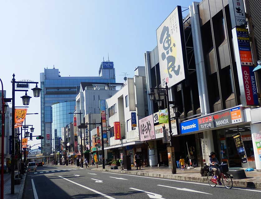A lively street in Hanno City, Japan, featuring local shops, colorful signs, and cyclists, representing community life and sustainable tourism development.