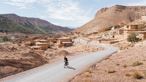 An aerial shot shows a lone cyclist riding on a winding paved road through a dry, mountainous desert landscape. Traditional clay-colored buildings of a village are clustered on the hillsides around the road. The sky is bright with some clouds.