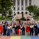A diverse group of approximately 30-40 people, smiling and waving with their hands raised, stand on a rainbow-painted crosswalk. Behind them, a large stately building with a dome (likely the Wisconsin State Capitol) is visible, flanked by trees and a statue.
