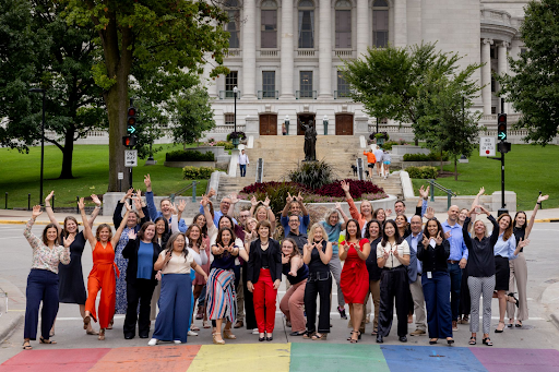 A diverse group of approximately 30-40 people, smiling and waving with their hands raised, stand on a rainbow-painted crosswalk. Behind them, a large stately building with a dome (likely the Wisconsin State Capitol) is visible, flanked by trees and a statue.