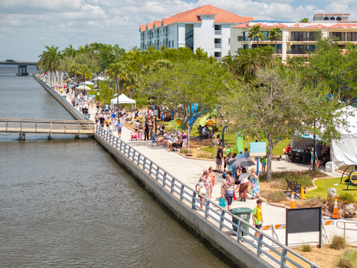 Visitors stroll along a lively waterfront promenade with trees, tents, and public art installations, showcasing a community placemaking event in a sunny coastal city.