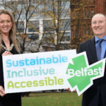 Two people standing outdoors in a city park hold a large sign that reads “Sustainable, Inclusive, Accessible Belfast.” Autumn trees and buildings appear in the background.