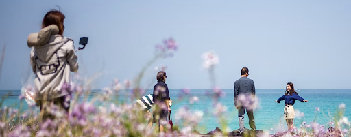 Visitors enjoy a scenic day by the turquoise waters of Jeju Island, surrounded by blooming flowers along the coast.