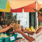A woman with a woven hat smiles as she receives a fresh pineapple from a vendor at an outdoor market stall.