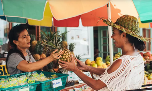 A woman with a woven hat smiles as she receives a fresh pineapple from a vendor at an outdoor market stall.