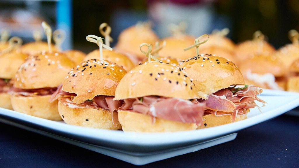 A plate of small prosciutto sliders with sesame seeds on buns, held together with toothpicks.