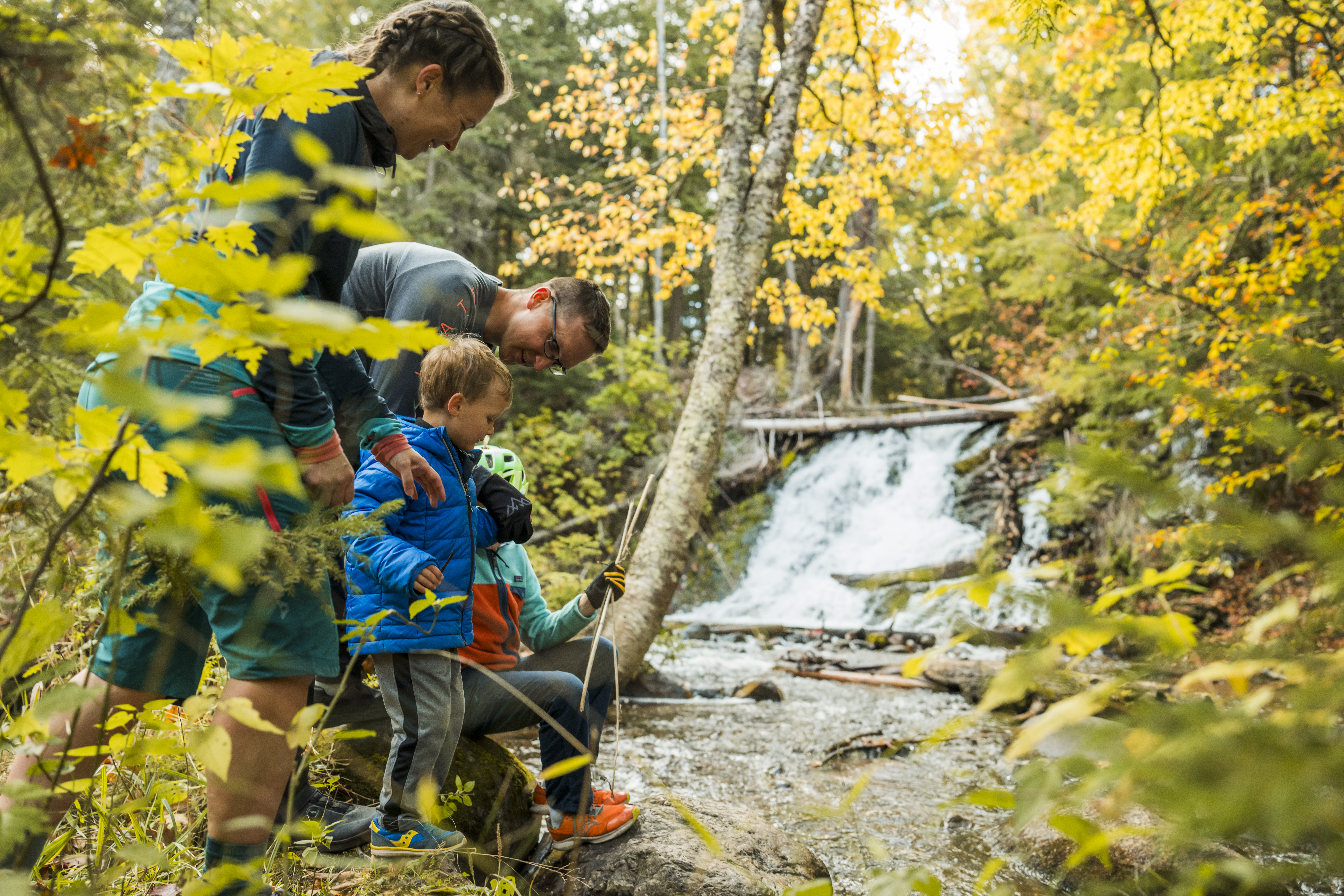 A family with two young children explores a forested trail beside a small waterfall in autumn, surrounded by yellow and green foliage. One adult kneels by the creek while the children play with sticks near the water.
