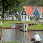 A cyclist rides along a canal in a quiet Amsterdam neighborhood, with traditional Dutch houses featuring red and blue facades visible across the water on a sunny day.