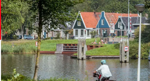 A cyclist rides along a canal in a quiet Amsterdam neighborhood, with traditional Dutch houses featuring red and blue facades visible across the water on a sunny day.
