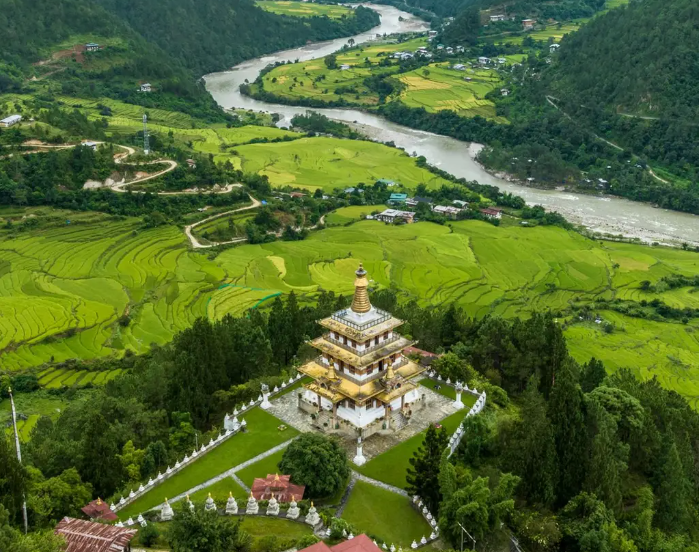 Aerial view of a traditional Bhutanese temple with a golden tiered roof, nestled in a lush green valley surrounded by rice terraces, forested hills, and scattered village homes.